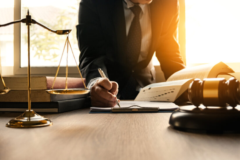 Lawyer writing on documents at a desk with a gavel, law books, and scales of justice